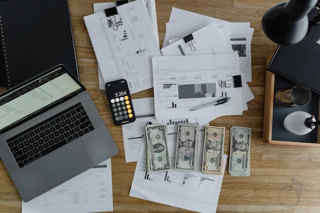 Desk covered with laptop, budget sheets, cash and a calculator.